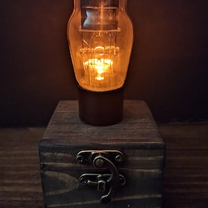 May include: A glowing vacuum tube lamp mounted on a dark, square wooden box. The clear glass bulb contains internal metal filaments emitting a warm orange light. The wooden base features a metallic latch mechanism on the front face.