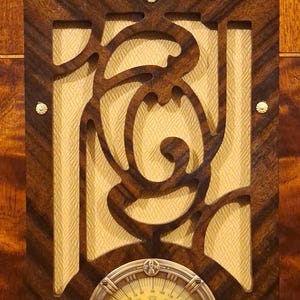 May include: Close-up of an antique radio with a dark wood veneer. The radio features a decorative, carved wooden overlay with a cream-colored fabric backing. A circular dial with a brass-colored bezel is at the bottom.