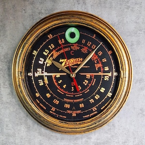 May include: A gold-colored clock face with black markings and a Zenith logo. The clock face has a dial with numbers 1 through 12 and a smaller dial with numbers 15 through 60. The clock face also has a red and gold hand.