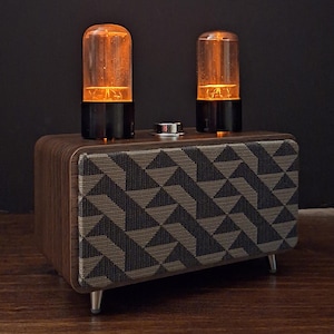 May include: A rectangular wooden speaker with a dark brown finish and a front grille featuring a geometric grey and black triangular pattern. Two glowing orange vacuum tubes are mounted on the top surface, alongside a central silver control knob.