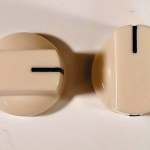 Puede incluir: Dos pomos de plástico de color crema con una línea negra en la parte superior. Los pomos son redondos con una sección central elevada.