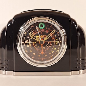 Puede incluir: Un reloj Zenith negro vintage con base plateada. La esfera presenta un diseño circular con marcas naranjas y negras, y agujas doradas. El reloj tiene un estilo retro, art déco.