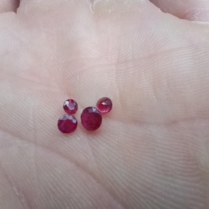 Op de afbeelding: Vier kleine, ronde, gefacetteerde edelstenen met een dieprode kleur. De stenen zijn in een groepje gerangschikt en vertonen een briljant slijpvorm met meerdere reflecterende oppervlakken die het licht vangen en hun levendige karmozijnrode tint tonen.