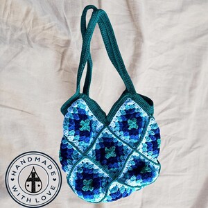 May include: A teal blue crocheted tote bag with a square pattern. The bag has two straps and a label that says "Handmade with Love" with a water drop and cross symbol.