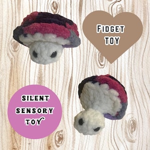 May include: A small, white, plush turtle with black eyes. The turtle's shell is made of a soft, fuzzy fabric in shades of pink, purple, and gray. The text "FIDGET TOY" is written on a brown heart-shaped background. The text "SILENT SENSORY TOY" is written on a pink circle.