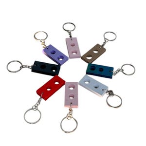 May include: Eight colorful keychains shaped like a two-stud Lego brick. Each keychain is a different color: red, pink, white, teal, black, brown, purple, and gray.