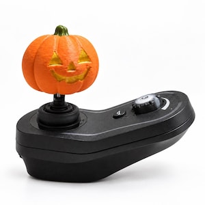 May include: A black electronic controller featuring a carved orange jack-o'-lantern pumpkin as the joystick topper. The base includes a circular dial with a power symbol, a small button with a symbol, and a curved ergonomic shape for handheld use.