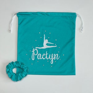 Puede incluir: Bolsa de cordón turquesa con un diseño de bailarina blanca y el nombre "Paetyn". Una scrunchie turquesa a juego está al lado. Pequeños adornos brillantes rodean el diseño.