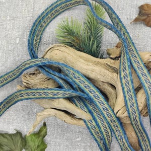 May include: Blue and tan woven ribbon with a geometric pattern. The ribbon is draped over driftwood and greenery, creating a natural and textured aesthetic. The ribbon's design features a repeating pattern of blue and tan stripes.