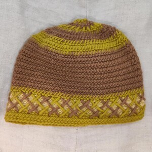 May include: A handmade crochet beanie in shades of brown and yellow. The hat features a textured stitch pattern with a decorative band of interwoven yellow and cream-colored yarn near the brim. The beanie is designed for warmth and style.