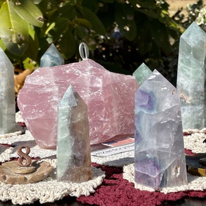 Puede incluir: Una colección de puntas de cristal verdes, moradas y blancas dispuestas en círculo sobre una mesa. Un gran cristal rosa se encuentra en el centro del círculo. Los cristales están sobre pequeñas bases de madera con números en ellas.
