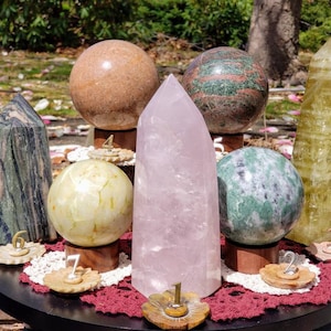 May include: A collection of seven polished stones, including spheres and points, arranged on a black circular surface with numbers 1 through 7. The stones are various shades of green, pink, brown, and yellow.