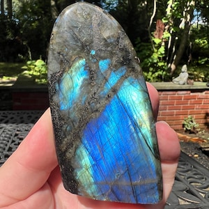 May include: A polished labradorite stone featuring a dark grey base with prominent iridescent blue and green flashes across its surface. The stone has a smooth, rounded top edge and a flat, irregular bottom edge with visible natural mineral inclusions.
