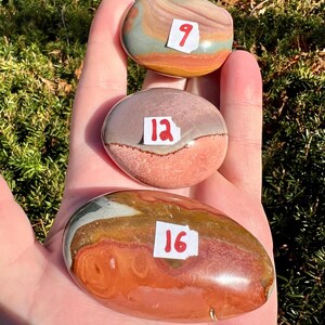 May include: Three polished stones in shades of brown, orange, pink, and green are held in a hand. Each stone has a white sticker with a red number: 9, 12, and 16. The stones have a smooth, rounded shape.