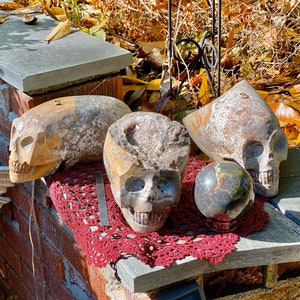 May include: Three carved stone skulls, one with a hole in the top, are arranged on a red crocheted doily with a black and gold sphere in the center.
