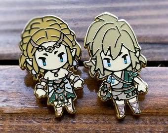 Op de afbeelding: Twee goudkleurige emaille pins met stripfiguren met blond haar en blauwe ogen. Eén personage draagt een groen-witte outfit en de ander een groen-beige outfit. De pins zijn geplaatst tegen een houten achtergrond.