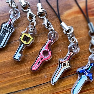 Op de afbeelding: Vijf acryl bedels met zilverkleurige sluitingen en zwarte koorden. De bedels hebben de vorm van zwaarden en een sleutel, met kleurrijke ontwerpen in zwart, roze, blauw en geel. De bedels liggen op een houten oppervlak.