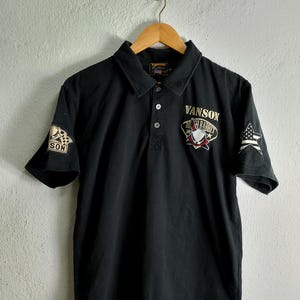 Può includere: Polo a maniche corte nero con un logo ricamato in oro che dice "Vanson Death Rabbit" sul davanti. La maglietta ha una toppa ricamata in oro sulla manica sinistra che dice "Vanson" e una toppa ricamata in oro sulla manica destra con un motivo a stella.