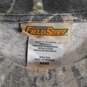 May include: Close-up of a gray camouflage-patterned t-shirt. The shirt has a tag with the brand name "Mossy Oak Field Staff" and text indicating it is made in Indonesia, 100% cotton, and size M/M/M.