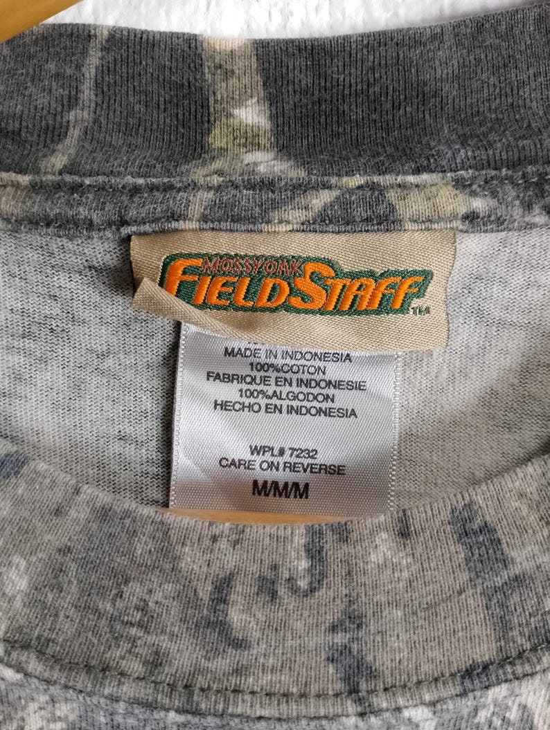 May include: Close-up of a gray camouflage-patterned t-shirt. The shirt has a tag with the brand name "Mossy Oak Field Staff" and text indicating it is made in Indonesia, 100% cotton, and size M/M/M.