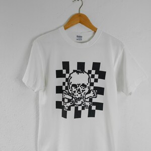 May include: White t-shirt with a black pixelated skull and crossbones design. The graphic is set on a checkered background. The t-shirt is displayed on a wooden hanger.