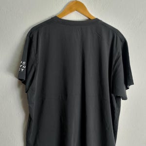 Puede incluir: Una camiseta gris oscuro colgada de una percha de madera. Se ve la parte trasera de la camiseta, con el texto "IS YOU CT" impreso verticalmente en la manga izquierda. La camiseta tiene cuello redondo y mangas cortas.