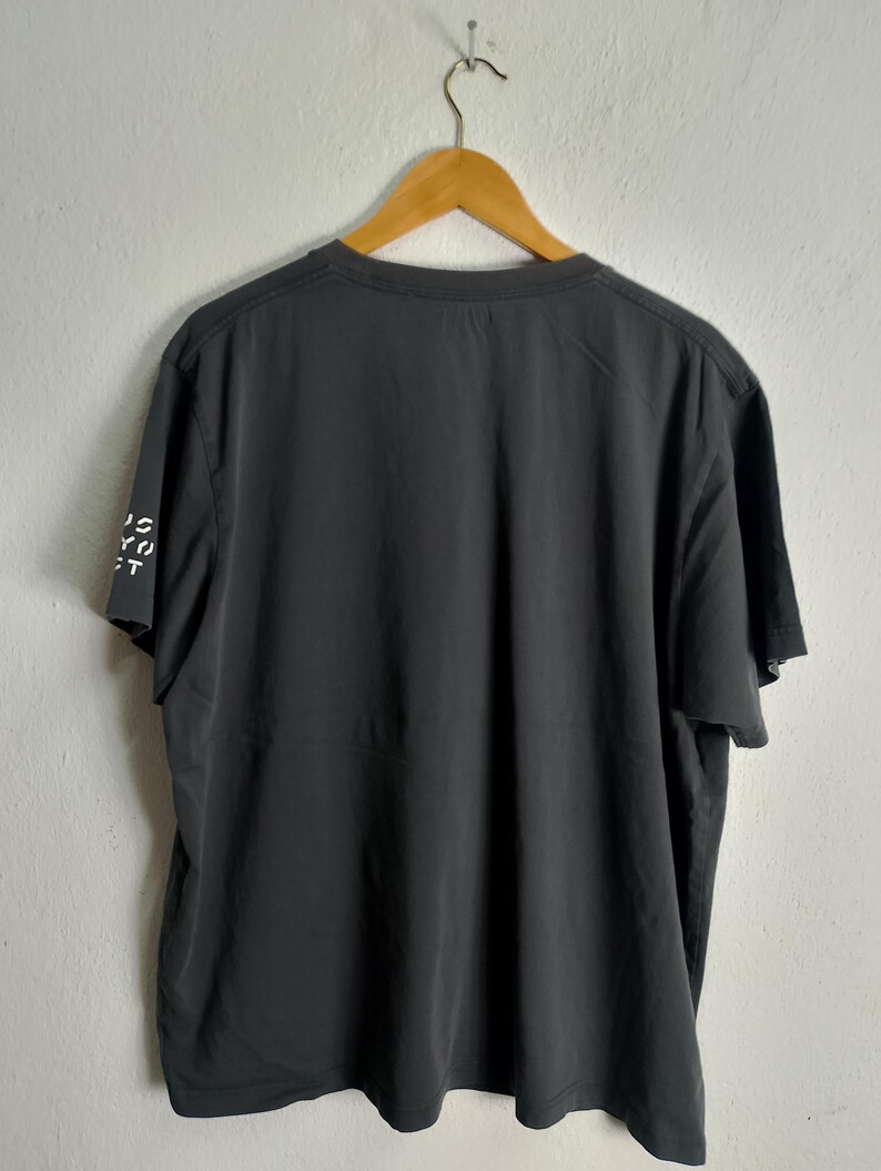 Puede incluir: Una camiseta gris oscuro colgada de una percha de madera. Se ve la parte trasera de la camiseta, con el texto "IS YOU CT" impreso verticalmente en la manga izquierda. La camiseta tiene cuello redondo y mangas cortas.