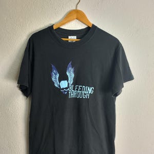 May include: Black t-shirt featuring a blue skull with feathered wings and the text "BLEEDING THROUGH" in a light blue font. The shirt is hanging on a wooden hanger against a neutral background.