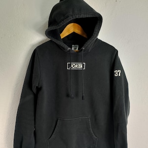 May include: Black hooded sweatshirt with a front pocket and drawstring. The word "JOKER" is printed in a white rectangular box on the chest. The number "37" is printed on the right sleeve. The sweatshirt is made of a soft material.