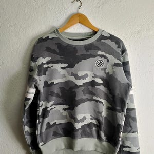 Könnte beinhalten: Ein graues Tarnmuster-Sweatshirt mit einem weißen Streifen an den Ärmeln und einem kleinen Logo auf der Vorderseite.