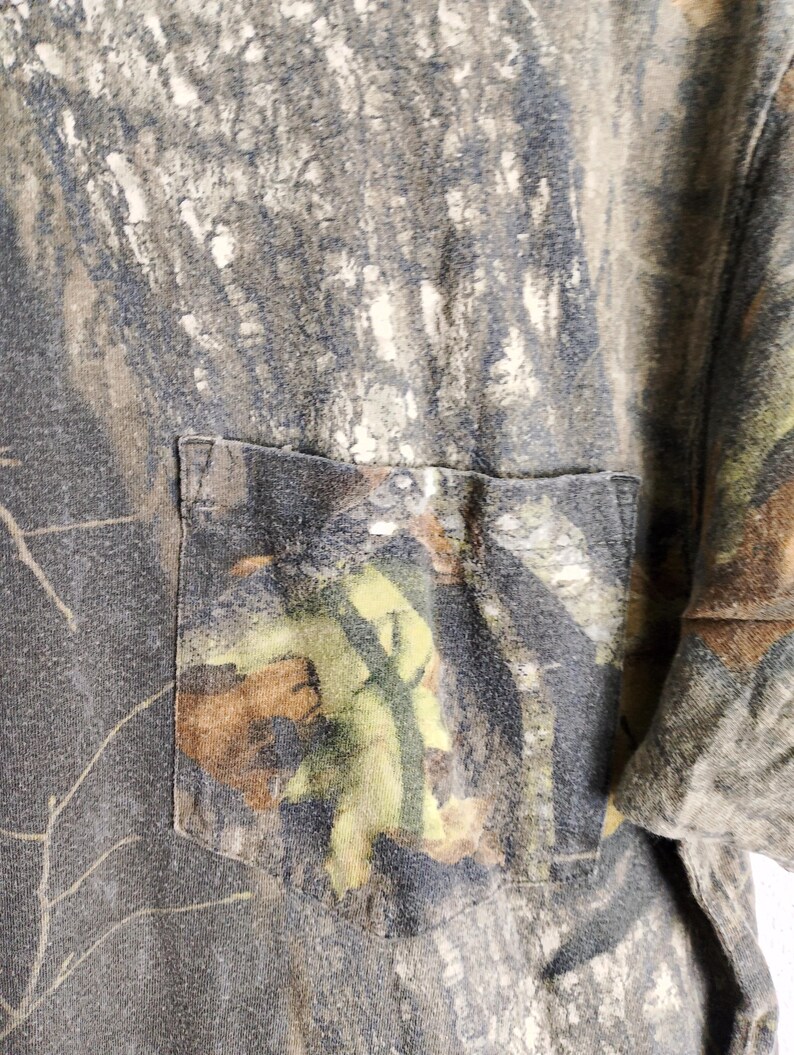 May include: Close-up of a camouflage-patterned t-shirt with a chest pocket. The shirt features a blend of greens, browns, and grays, mimicking a forest environment. The pocket is a darker shade of the same camouflage print.