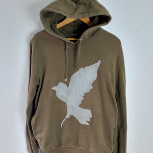 Può includere: Felpa con cappuccio verde oliva con una grande grafica a colomba grigio chiaro strutturata sul davanti. La felpa ha un cappuccio con coulisse e maniche lunghe. L'indumento è esposto su una gruccia di legno.