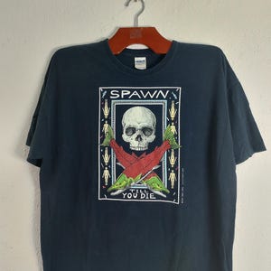 May include: Dark navy blue t-shirt with a graphic design featuring a skull, crossed fish, and the text "SPAWN" and "TILL YOU DIE". The design is framed in a white rectangle with small figures.