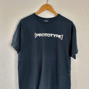 Puede incluir: Una camiseta azul marino con la palabra "PROTOTYPE" impresa en blanco, exhibida en una percha de madera. El texto está en negrita, sin serifa, con un efecto ligeramente desgastado. La camiseta cuelga contra una pared lisa de color blanquecino.