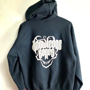 Puede incluir: Sudadera con capucha azul marino con el texto "Knowledge Bones" en una fuente blanca y estilizada. El diseño presenta un gráfico de calavera. La sudadera tiene una capucha con cordón y un bolsillo de canguro.