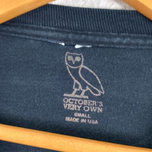 Pode incluir: Close-up de uma camiseta preta com um logotipo de coruja branca e texto. O texto diz "OCTOBER'S VERY OWN" e "SMALL MADE IN USA". A camisa est&aacute; pendurada em um cabide de madeira.