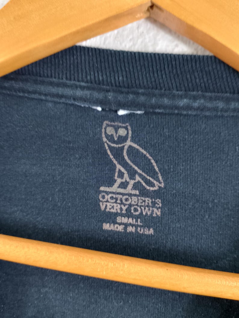 Pode incluir: Close-up de uma camiseta preta com um logotipo de coruja branca e texto. O texto diz "OCTOBER'S VERY OWN" e "SMALL MADE IN USA". A camisa est&aacute; pendurada em um cabide de madeira.