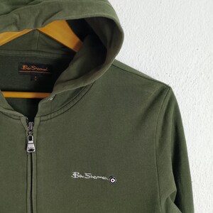 May include: Olive green zip-up hoodie with a hood and white paneling. The Ben Sherman logo is embroidered on the chest. The hoodie is made of a soft, comfortable material.
