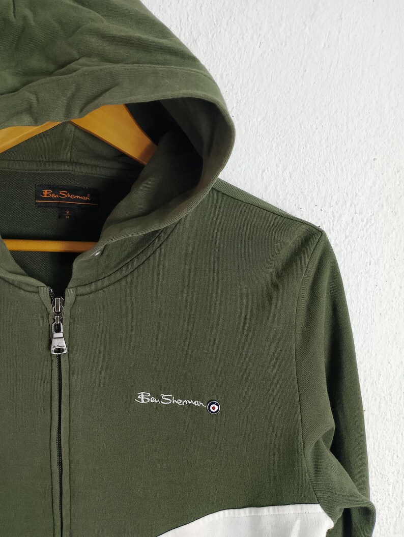 May include: Olive green zip-up hoodie with a hood and white paneling. The Ben Sherman logo is embroidered on the chest. The hoodie is made of a soft, comfortable material.