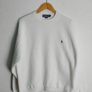 May include: A white crew neck sweatshirt with the Ralph Lauren logo embroidered in navy blue on the chest. The sweatshirt is hanging on a wooden hanger against a white wall. The Ralph Lauren label is visible at the neckline.