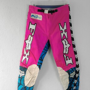May include: Bright pink motocross pants with white and blue accents. The pants feature the "FOX" logo in white on the sides and a black belt. The lower legs have a blue and white geometric pattern.