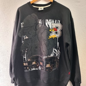 Könnte beinhalten: Schwarzes Sweatshirt mit einem grafischen Design, das eine Zeichentrickfigur mit Baseballkappe, den Buchstaben "B" und zwei schwarzen Trucks zeigt. Der Text "School Of Hard Knocks" ist ebenfalls sichtbar.