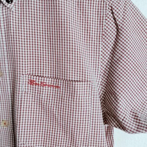 K&ouml;nnte beinhalten: Ein kurz&auml;rmeliges Hemd mit Knopfleiste und einem rot-wei&szlig;en Gingham-Karomuster. Das Hemd verf&uuml;gt &uuml;ber eine Brusttasche mit dem in rotem Garn gestickten Schriftzug Ben Sherman dar&uuml;ber sowie helle Kn&ouml;pfe entlang der vorderen Knopfleiste.
