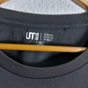 Puede incluir: Camiseta gris oscuro de cuello redondo con los logotipos "UT" y "KAWS TOKYO FIRST" estampados. La camiseta es de talla 3XL y est&aacute; fabricada en China. Cuelga de una percha de madera.