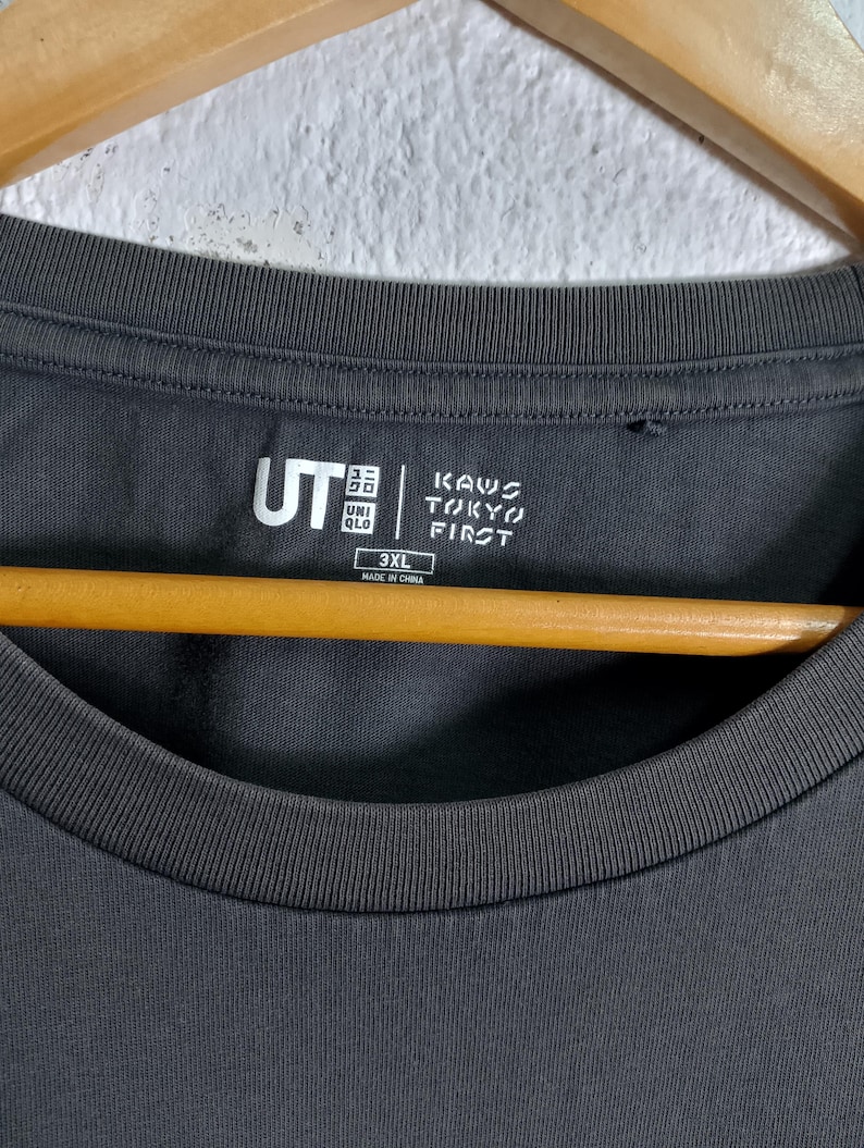 Puede incluir: Camiseta gris oscuro de cuello redondo con los logotipos "UT" y "KAWS TOKYO FIRST" estampados. La camiseta es de talla 3XL y est&aacute; fabricada en China. Cuelga de una percha de madera.