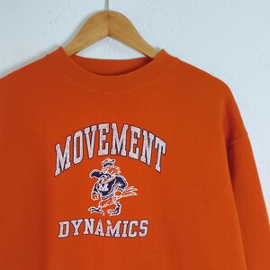 以下が含まれることがあります： オレンジ色のスウェットシャツ。白文字で「MOVEMENT DYNAMICS」と書かれています。「DYNAMICS」という単語の上に、船乗りの帽子をかぶった漫画のマスコットが描かれています。スウェットシャツは木製のハンガーに吊るされています。