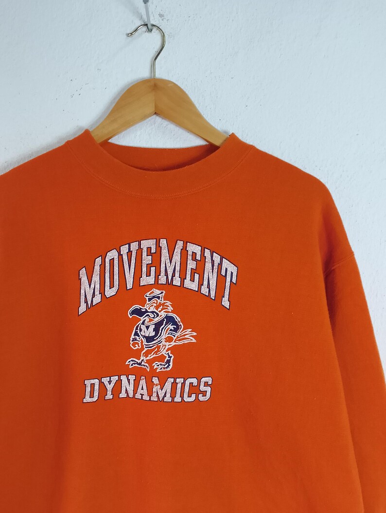以下が含まれることがあります： オレンジ色のスウェットシャツ。白文字で「MOVEMENT DYNAMICS」と書かれています。「DYNAMICS」という単語の上に、船乗りの帽子をかぶった漫画のマスコットが描かれています。スウェットシャツは木製のハンガーに吊るされています。