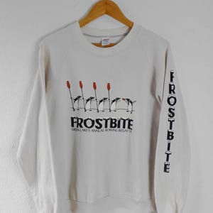 Puede incluir: Sudadera de color crema con la palabra "FROSTBITE" en negro y el texto "GREEN LAKES ANNUAL ROWING REGATTA" debajo. La manga tiene la palabra "FROSTBITE" impresa verticalmente. El diseño presenta pingüinos y remos rojos.