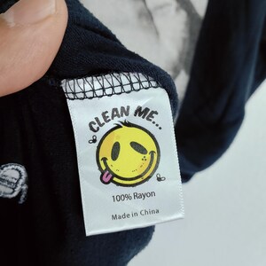 May include: Close-up of a navy blue garment tag with the text "CLEAN ME..." above a yellow smiley face with its tongue out. The tag also reads "100% Rayon" and "Made in China".