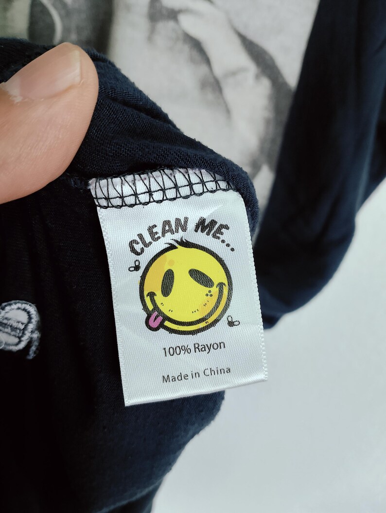 May include: Close-up of a navy blue garment tag with the text "CLEAN ME..." above a yellow smiley face with its tongue out. The tag also reads "100% Rayon" and "Made in China".